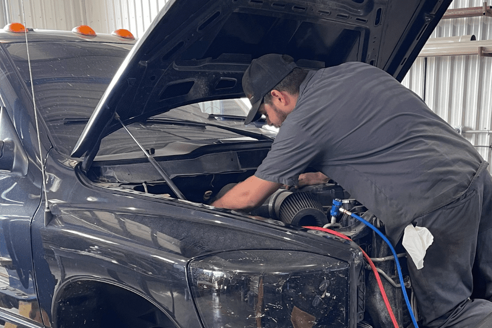 Trusted diesel mechanics in Palmerston, ON at CDP Diesel & Auto. A mechanic performing diagnostics under the hood of a black diesel truck, with A/C service hoses connected. Captures hands-on diesel repair service in a well-lit shop environment.